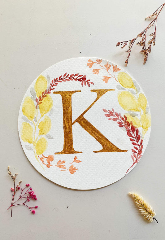 Round watercolor monogram of the letter K painted in metallic gold watercolor, surrounded by deep red, orange and yellow watercolor leaves on a white surface flanked with dried flowers.