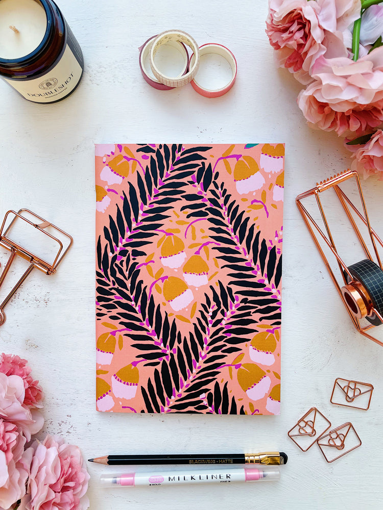 Flat Lay Photograph of Peach Poetry A5 Notebook Featuring Black Leaves on Intertwining Branches with Semi-Abstract Flowers in Light Brown on a Peach Pink Background Surrounded by Pink Flowers and Stationery Supplies.
