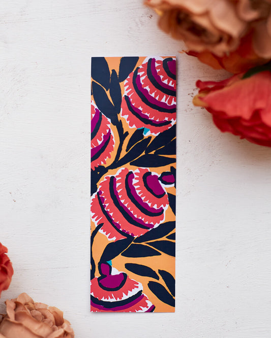 A glossy 2 x 6 bookmark depicting  interlacing branches adorned with leaves in black with large, semi-abstract flowers in red, purple, white, and black on a mustard yellow ground.