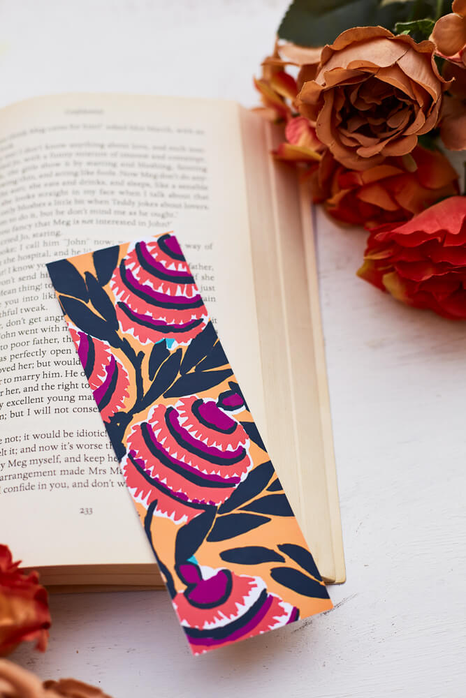 A glossy 2 x 6 bookmark depicting  interlacing branches adorned with leaves in black with large, semi-abstract flowers in red, purple, white, and black on a mustard yellow ground on an open book surrounded by flowers.