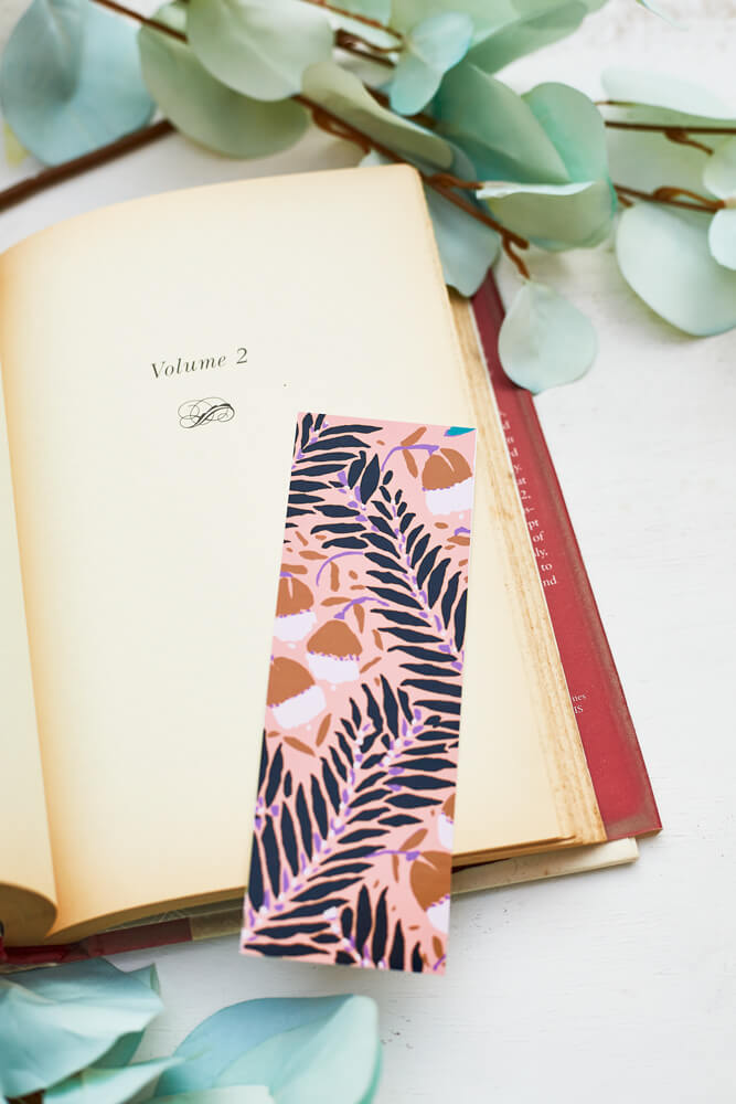 A glossy 2 x 6 bookmark depicting scrolling branches, abstract flowers, and a peachy pink background on an open book with leaves in the background.