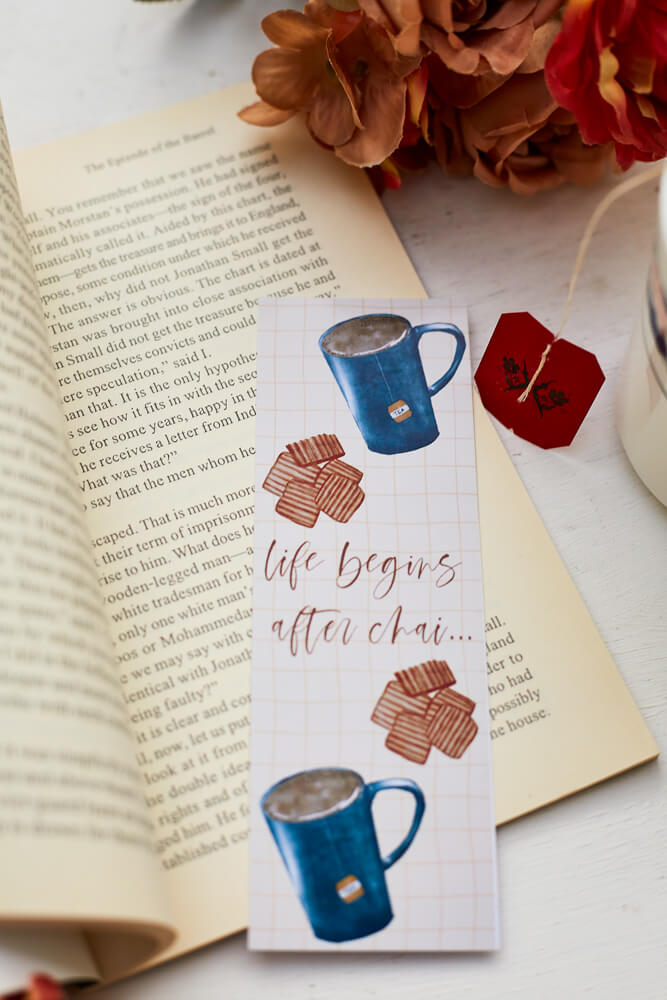 A glossy 2 X 6 bookmark with a pale yellow grid background depicting large mugs of tea and biscuits with the words "life begins after chai..." in script lettering across the middle on an open book next to a cup of tea with orange and brown flowers in the background.
