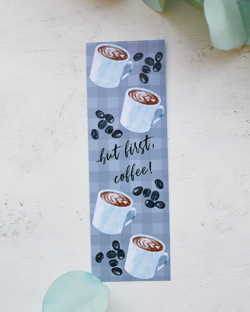 A glossy 2 X 6 bookmark with a pale grey plaid background, depicting cups of coffee and coffee beans with the words “…but first, coffee!” in script lettering across the middle.