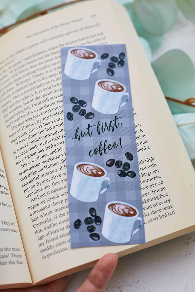 A glossy 2 X 6 bookmark with a pale grey plaid background, depicting cups of coffee and coffee beans with the words “…but first, coffee!” in script lettering across the middle on an open book with leaves in the background.