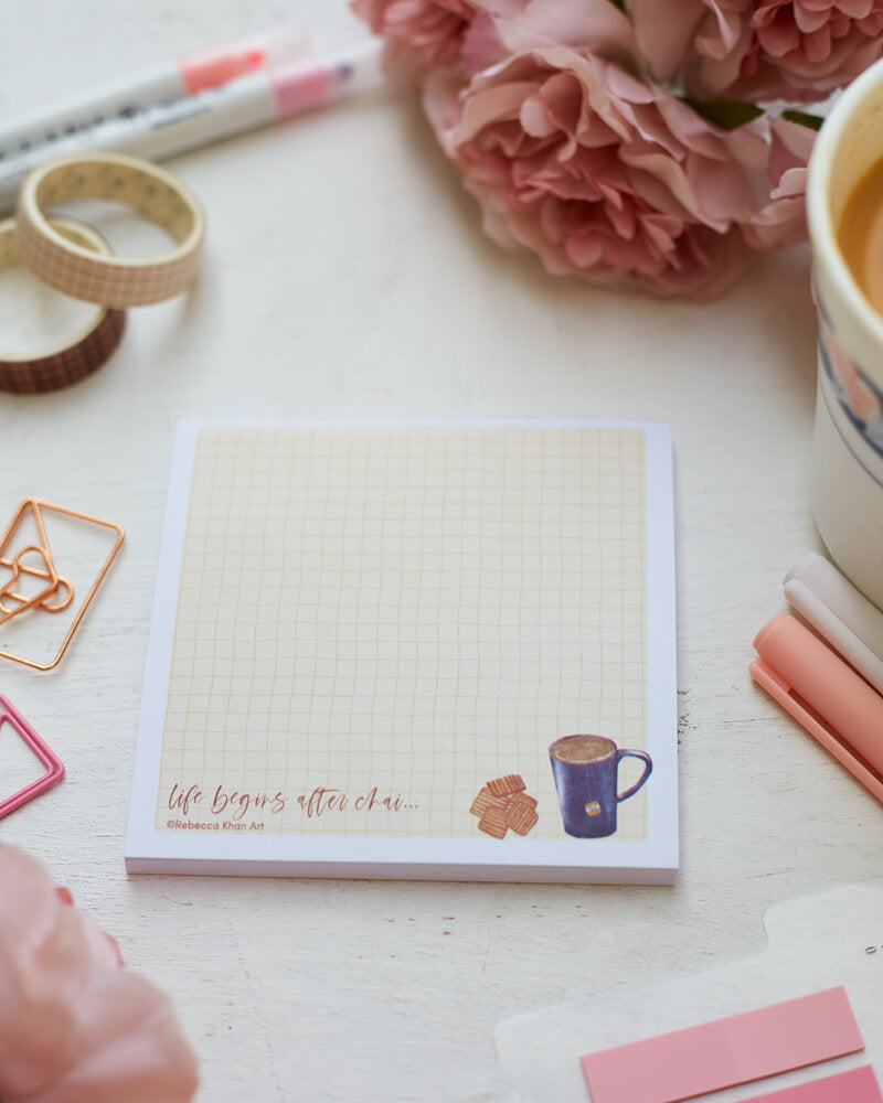 A notepad with a pale yellow grid background with a white border, on the bottom a cup of tea and biscuits are depicted with the words "life begins after chai..." in script lettering. The notepad is on a desk surrounded by pink flowers and stationery supplies as well as a cup of tea.