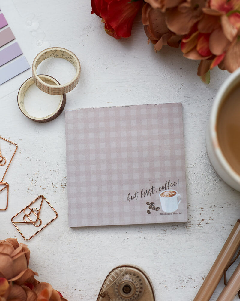 A notepad with a pale grey-brown plaid background, on the bottom of which a cup of coffee and some coffee beans are depicted with the words “…but first, coffee!” in script lettering. The notepad is on a desk surrounded by orange and brown flowers and stationery supplies as well as a cup of coffee.