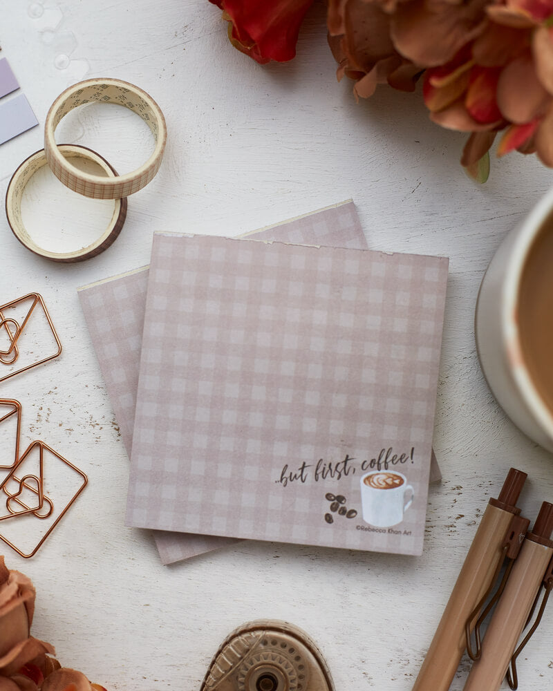 Two notepads with a pale grey-brown plaid background, on the bottom of which a cup of coffee and some coffee beans are depicted with the words “…but first, coffee!” in script lettering. The notepads are on a desk surrounded by orange and brown flowers and stationery supplies as well as a cup of coffee.