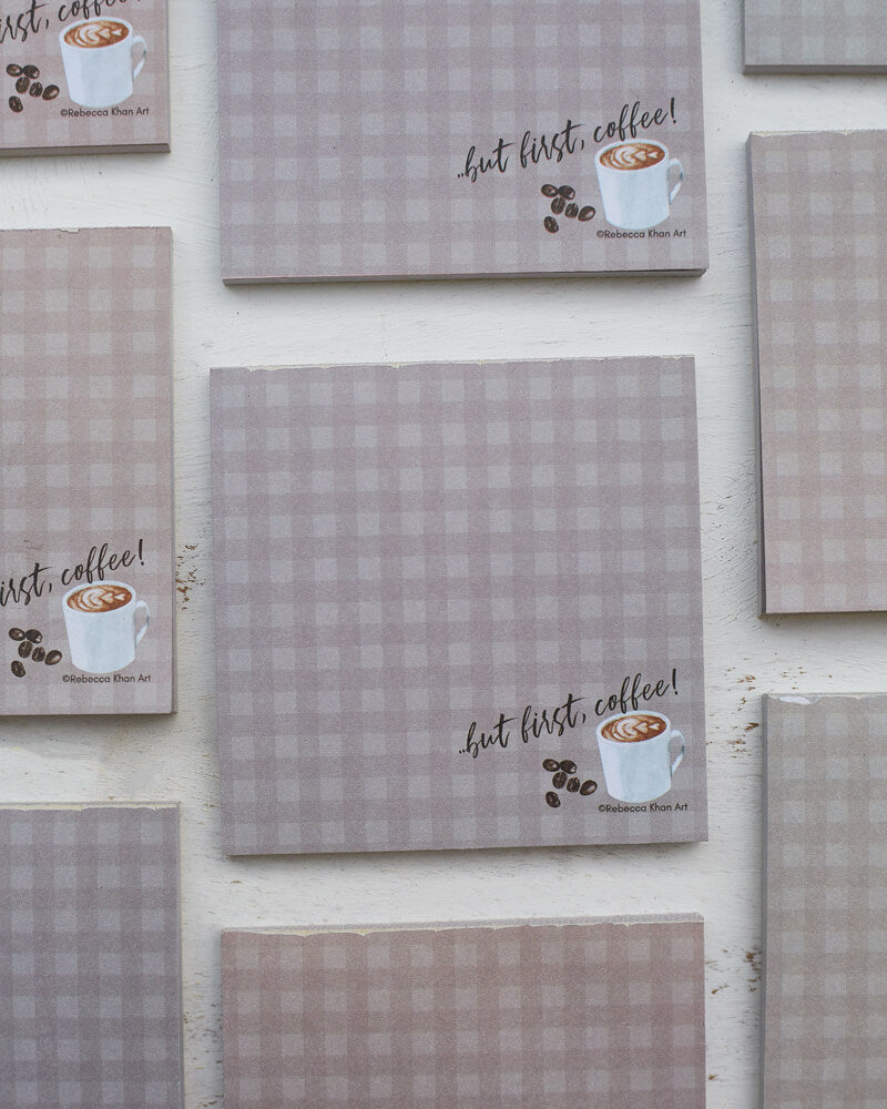 Multiple notepads with a pale grey-brown plaid background, on the bottom of which a cup of coffee and some coffee beans are depicted with the words “…but first, coffee!” in script lettering.