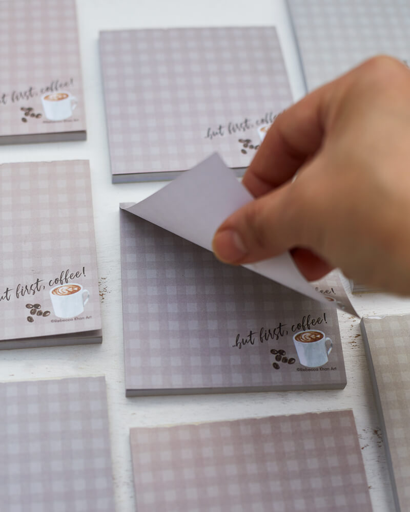 Close up of a hand tearing off a note from a notepad with a pale grey-brown plaid background, on the bottom of which a cup of coffee and some coffee beans are depicted with the words “…but first, coffee!” in script lettering.