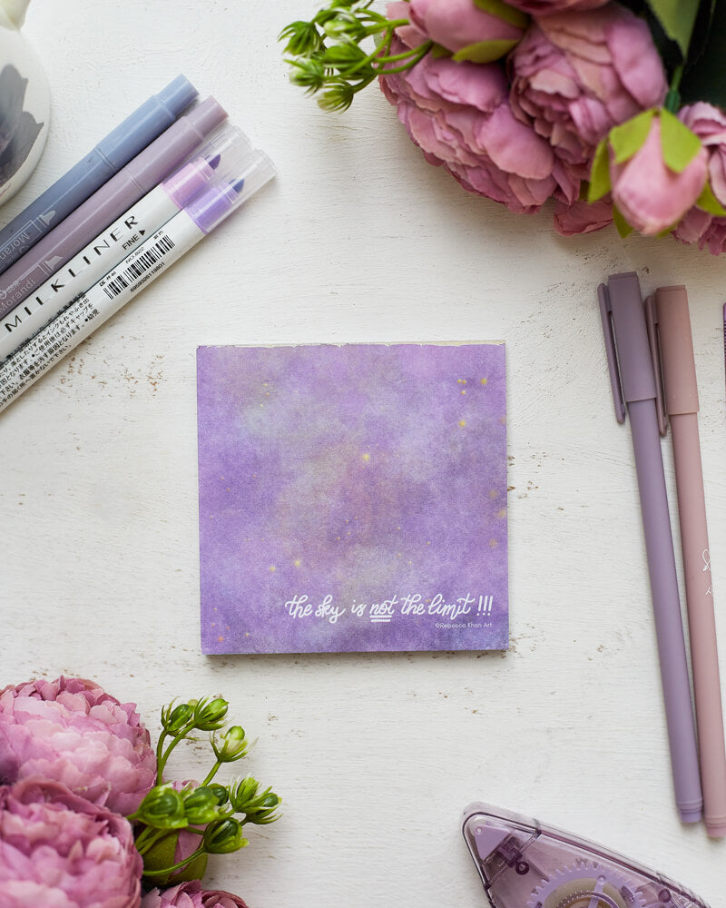 A notepad depicting a starry galaxy against a light purple background with the words "The sky is NOT the limit!!!" in white hand lettering across the bottom.  The notepad are on a desk surrounded by flowers and purple stationery supplies.