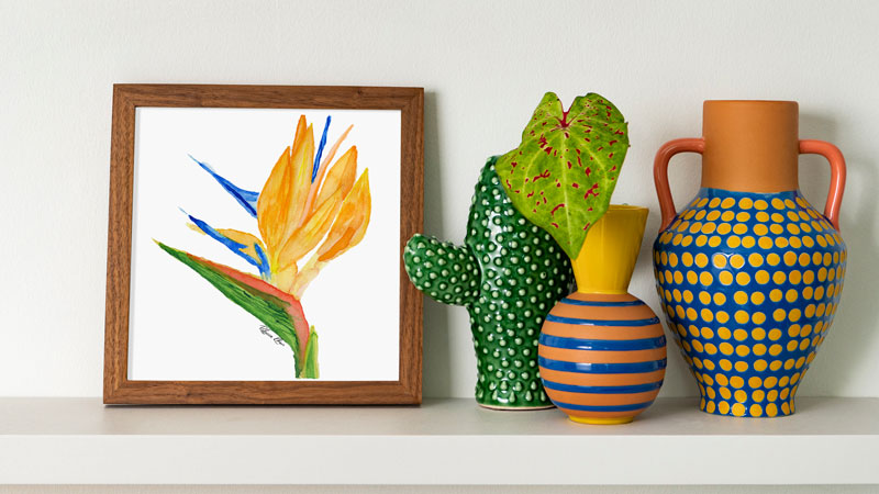 An art print of a watercolor painting of a bird of paradise flower.  The art print is in a square wooden frame on a shelf next to some small colorful vases.