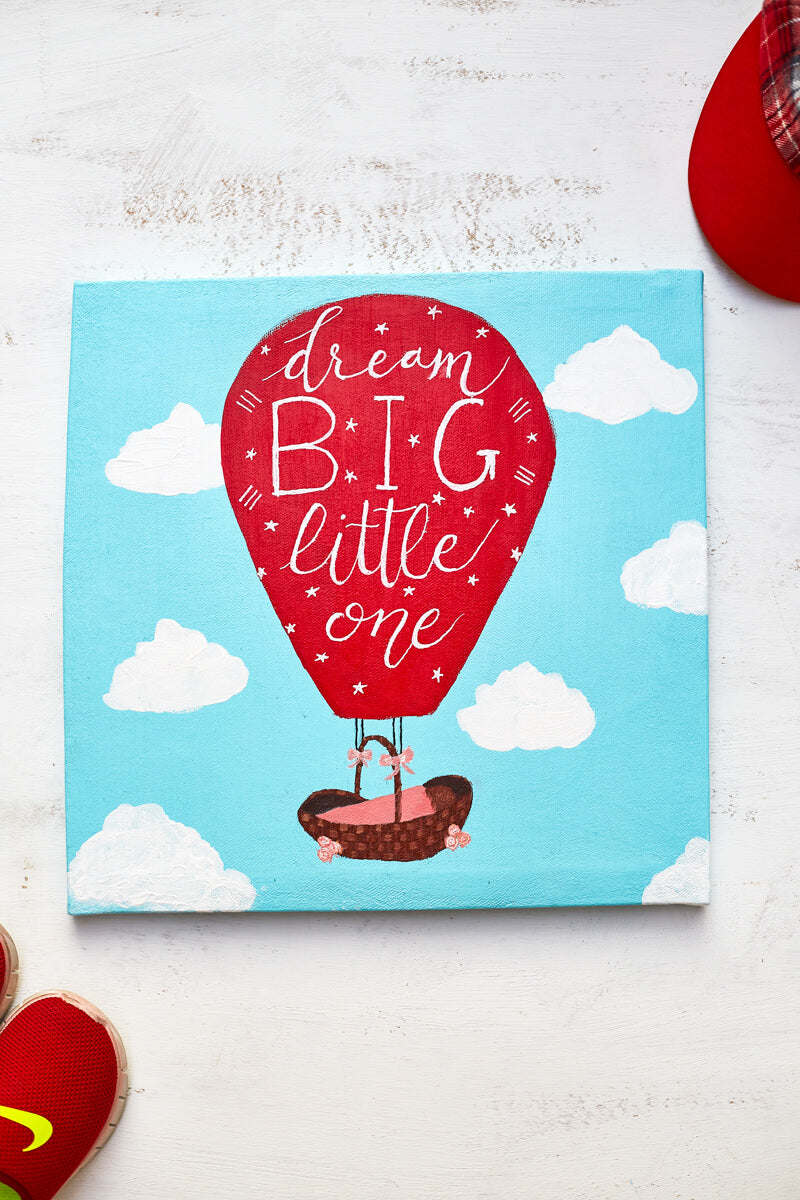 Acrylic painting on canvas of a bright red hot air balloon featuring the words "Dream Big Little One" in hand lettering ascending the sky with a baby basket.  The painting is surrounded by a bright red child's cap and red shoes.