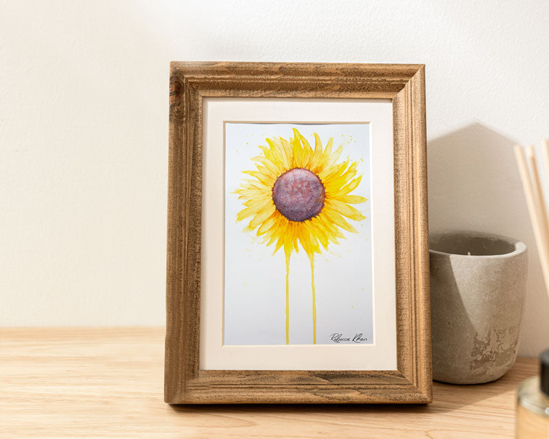 Art print of a watercolor painting of a large sunflower with petals radiating outward and two petals dripping down to the bottom of the painting inside a wooden frame on a table next to a candle.