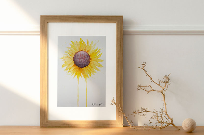 Art print of a watercolor painting of a large sunflower with petals radiating outward and two petals dripping down to the bottom of the painting in a wooden frame on a table with decoration pieces next to it.