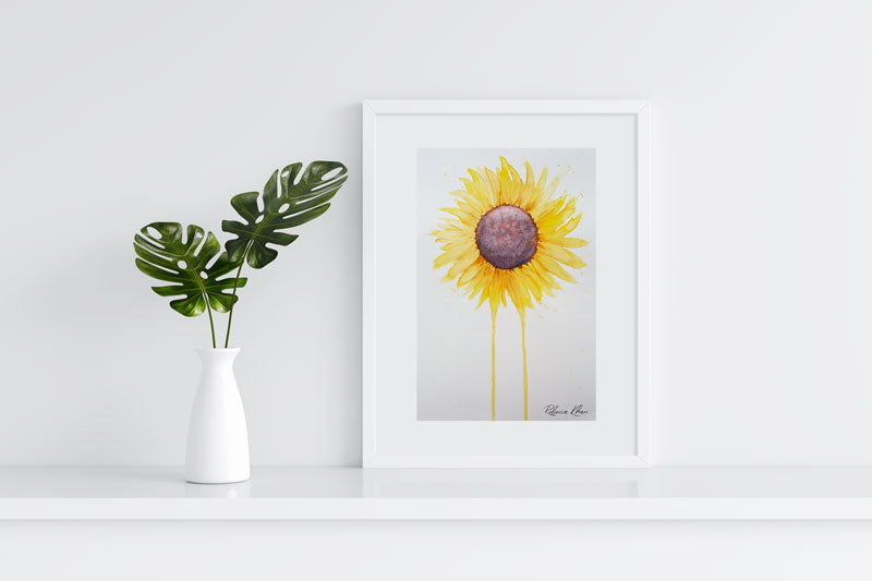 Art print of a watercolor painting of a large sunflower with petals radiating outward and two petals dripping down to the bottom of the painting in a white frame on a white shelf next to a white vase with two leaves in it.