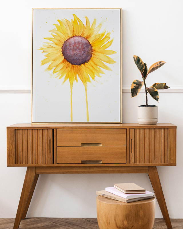 A side table with a plant and a large watercolor painting of a  sunflower with petals radiating outward and two petals dripping down to the bottom of the painting.