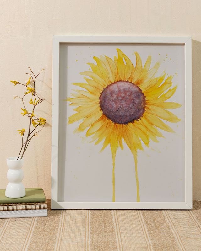 A large watercolor painting of a sunflower with petals radiating outward and two petals dripping down to the bottom of the painting in a white frame against a wall next to books and a vase with dried flowers.