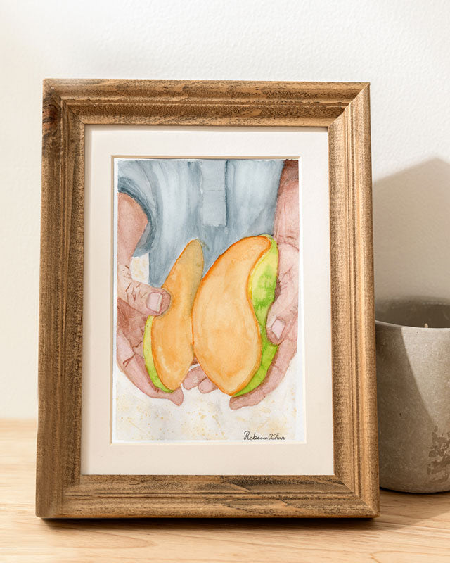 A wooden frame with an art print of a watercolor painting of two hands holding a bright mango that has been cut into to reveal the inside.