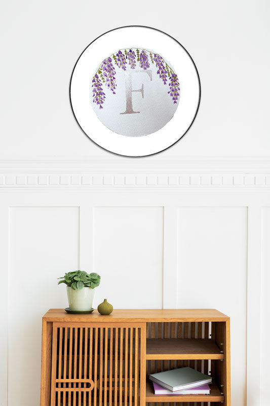 Watercolor monogram of the letter F in silver watercolor with purple watercolor wisteria hanging down around it on a round piece of paper in a round frame above a side table.