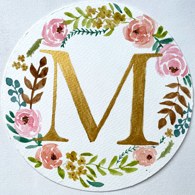 Watercolor monogram of the letter M in gold watercolor surrounded by pink and peach watercolor peonies and green and brown leaves on a round piece of paper.