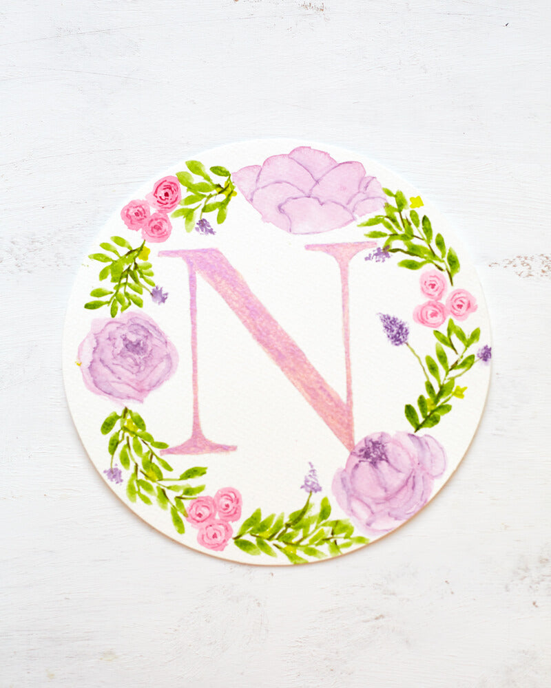 Round watercolor monogram painting of the letter N painted in duochrome pink and purple metallic paint surrounded by painted purple peonies and pink roses and green leaves.