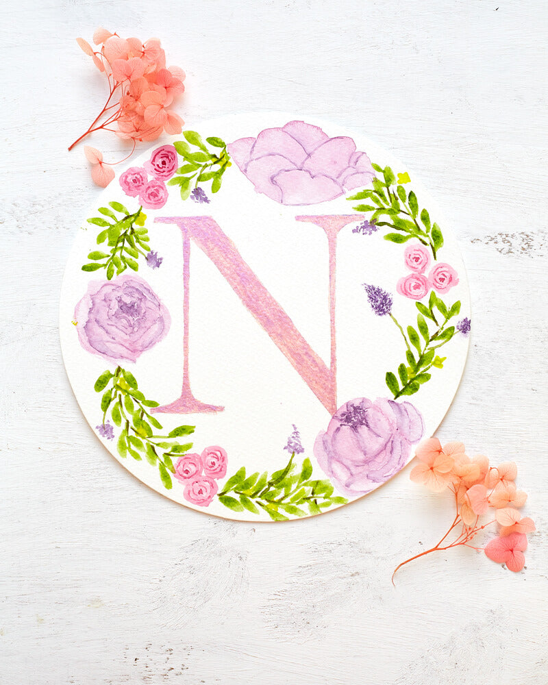 Round watercolor monogram painting of the letter N painted in duochrome pink and purple metallic paint surrounded by painted purple peonies and pink roses and green leaves flanked by dried pink flowers.
