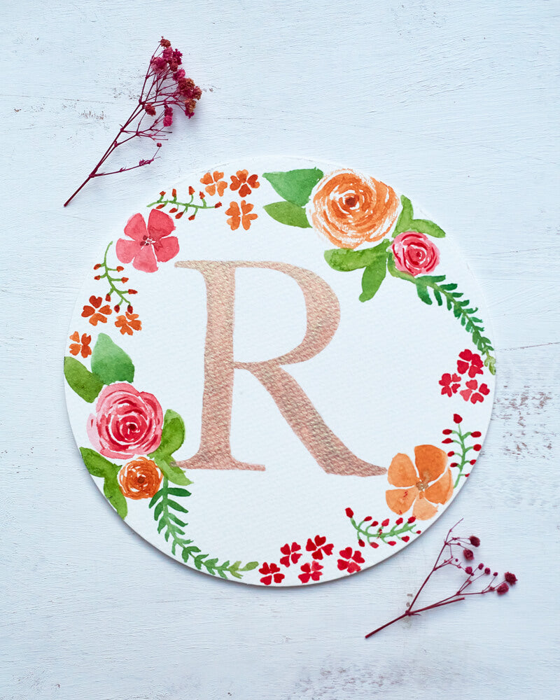 Two dried red flowers flanking a watercolor monogram of the letter R painted in metallic duochrome peach and green paint with red and orange flowers and green leaves painted around it on a round paper.