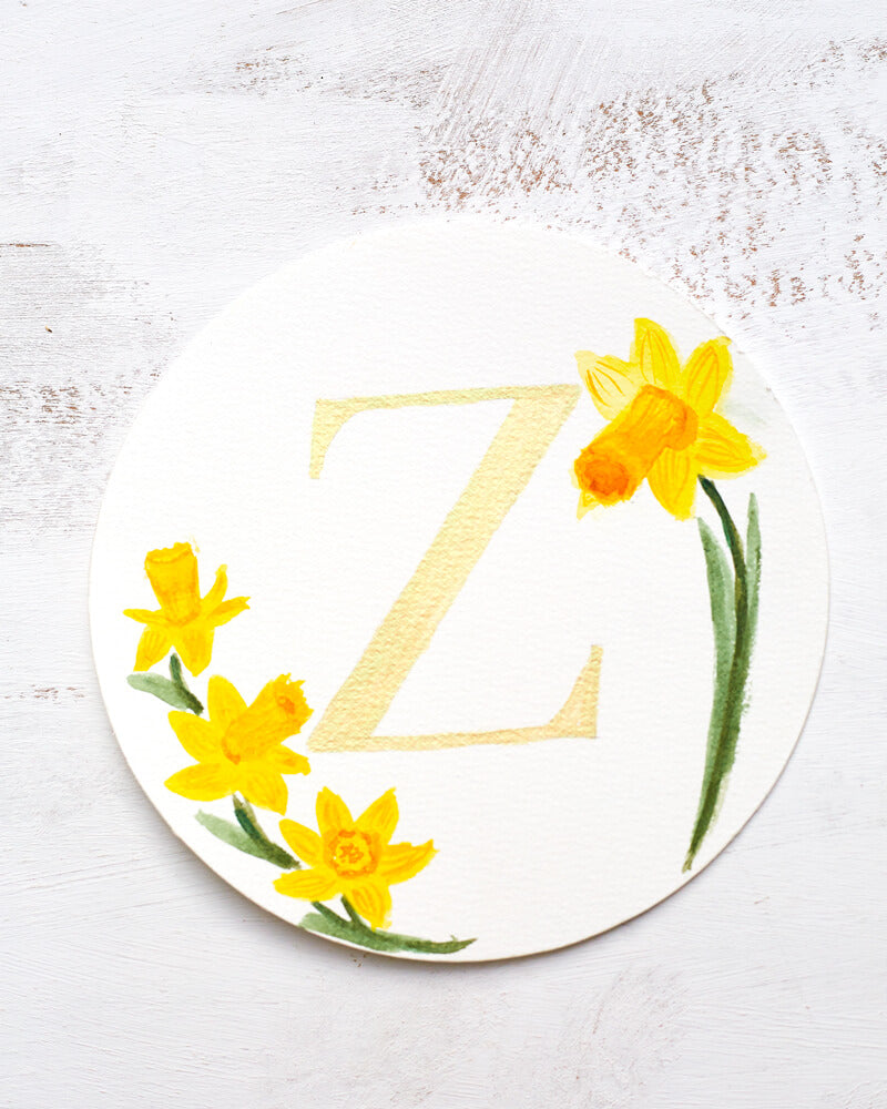 Watercolor monogram of the letter Z painted in duochrome metallic paint with pale gold and green shine surrounded by painted daffodils on a round piece of paper.