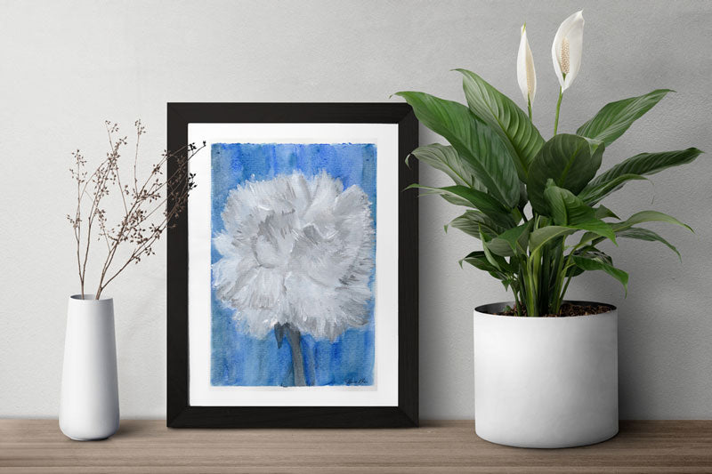Art print of a  mix media abstract floral painting of a white carnation on a blue background in a black frame on a table in between two plants.