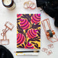 Flat lay photo of the Amber Harmony A5 Size Notebook with Bright Colorful Flowers on A Yellow Background Surrounded by Stationery Supplies and a Black Scarf.