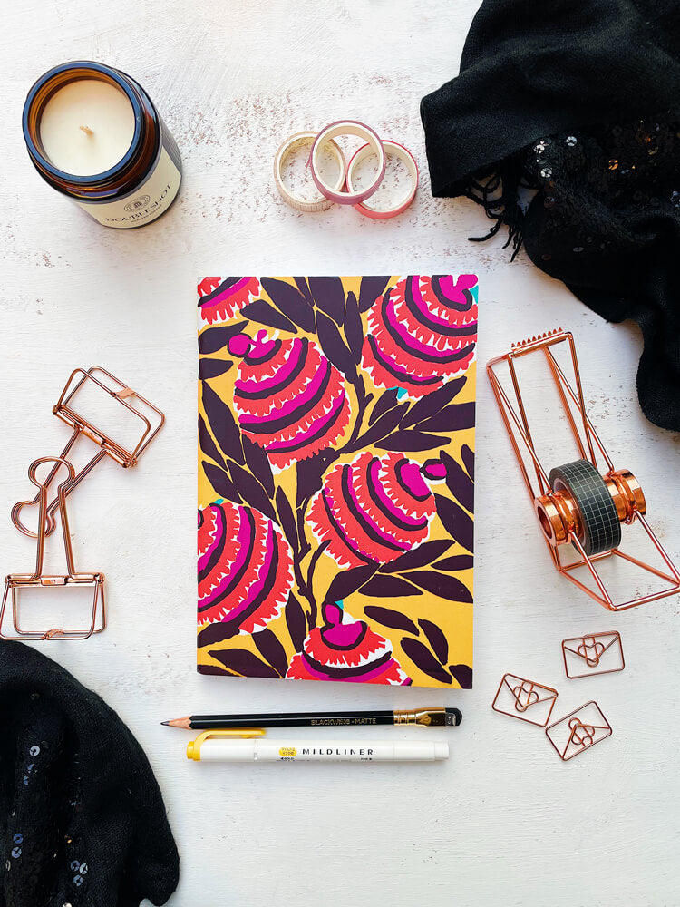 Flat lay photo of the Amber Harmony A5 Size Notebook with Bright Colorful Flowers on A Yellow Background Surrounded by Stationery Supplies and a Black Scarf.