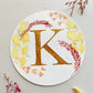 Round watercolor monogram of the letter K painted in metallic gold watercolor, surrounded by deep red, orange and yellow watercolor leaves on a white surface flanked with dried flowers.