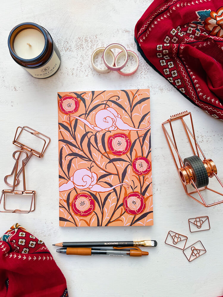 Flat Lay Photograph of the Oriental Elegance Softcover A5 Notebook  Featuring Intertwining Branches with Scrolling Leaves with Abstract Oriental Poppies and Abstract Clouds on a Sand-Colored Background Surrounded by Stationery Items a Candle and a Red Scarf.