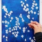 An acrylic painting on canvas depicting white blossoms against a moody blue background with a hand holding a paintbrush against it.