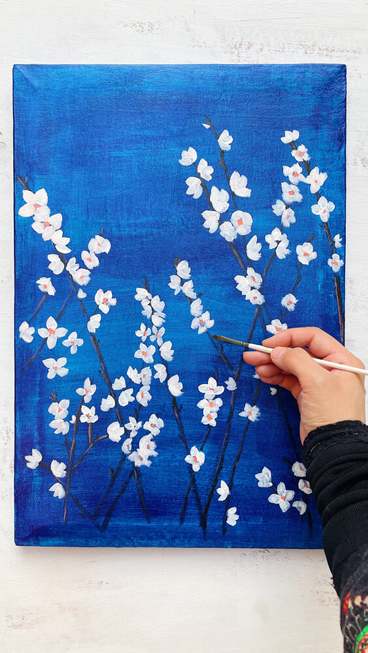 An acrylic painting on canvas depicting white blossoms against a moody blue background with a hand holding a paintbrush against it.