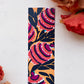 A glossy 2 x 6 bookmark depicting  interlacing branches adorned with leaves in black with large, semi-abstract flowers in red, purple, white, and black on a mustard yellow ground.