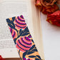 A glossy 2 x 6 bookmark depicting  interlacing branches adorned with leaves in black with large, semi-abstract flowers in red, purple, white, and black on a mustard yellow ground on an open book surrounded by flowers.