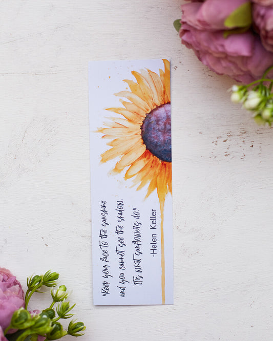 A glossy 2 x 6 inch bookmark depicting a yellow sunflower painted in watercolor with the lower petals dripping.  On the side is written, "Keep your face to the sunshine and you cannot see the shadow.  It's what sunflowers do. -Helen Keller."