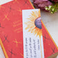 A glossy 2 x 6 inch bookmark depicting a yellow sunflower painted in watercolor with the lower petals dripping.  On the side is written, "Keep your face to the sunshine and you cannot see the shadow.  It's what sunflowers do. -Helen Keller."  The bookmark is resting on a closed book and in the background are purple flowers.