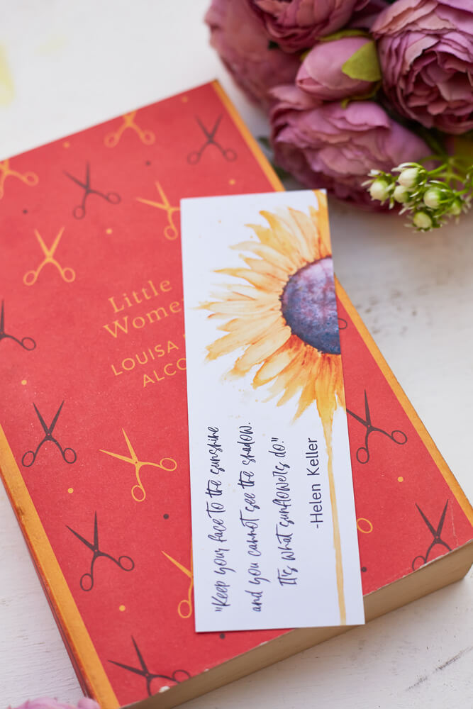 A glossy 2 x 6 inch bookmark depicting a yellow sunflower painted in watercolor with the lower petals dripping.  On the side is written, "Keep your face to the sunshine and you cannot see the shadow.  It's what sunflowers do. -Helen Keller."  The bookmark is resting on a closed book and in the background are purple flowers.