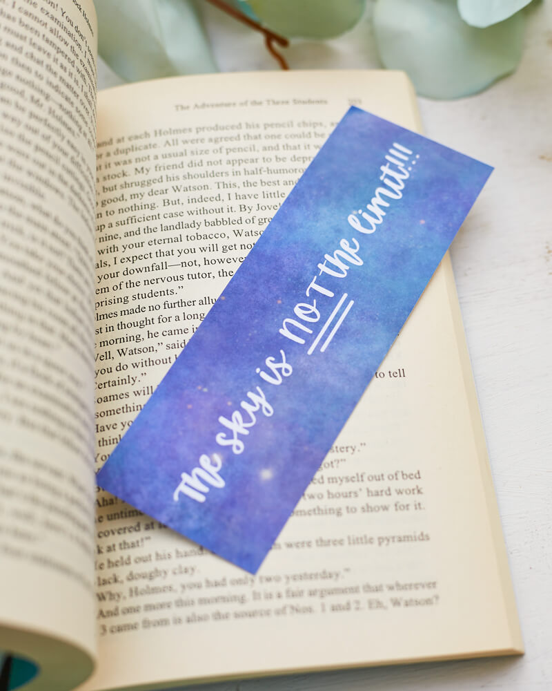 A glossy 2 x 6 bookmark depicting a starry galaxy against a light purple background with the words "The sky is NOT the limit!!!" in white hand lettering across the middle on an open book with leaves in the background.