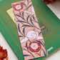 A glossy 2 x 6 inch bookmark depicting delicate branches, scrolling leaves, and semi-abstract flowers on top of a green book surrounded by orange and brown flowers.