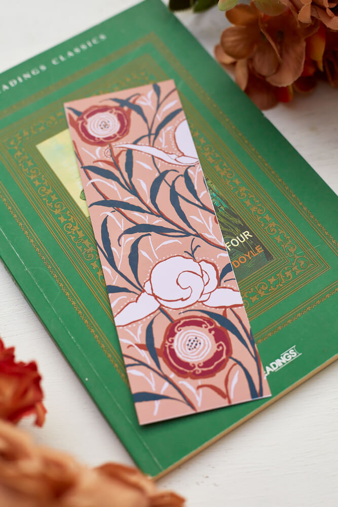 A glossy 2 x 6 inch bookmark depicting delicate branches, scrolling leaves, and semi-abstract flowers on top of a green book surrounded by orange and brown flowers.