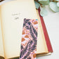 A glossy 2 x 6 bookmark depicting scrolling branches, abstract flowers, and a peachy pink background on an open book with leaves in the background.