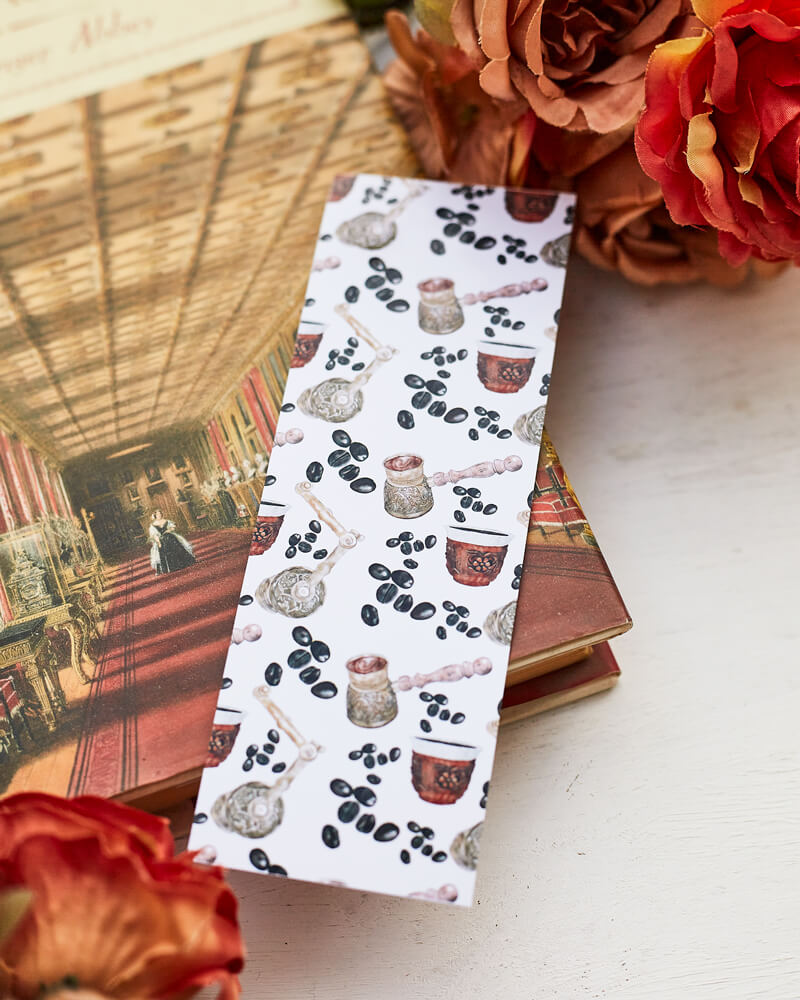 A glossy 2 X 6 inch bookmark depicting a pattern of coffee beans, cups of turkish coffee, a Turkish coffee grinder and a cezve.  The bookmark rests on the cover of a book, surrounded by orange and brown flowers.