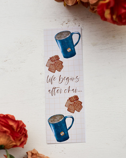 A glossy 2 X 6 bookmark with a pale yellow grid background depicting large mugs of tea and biscuits with the words "life begins after chai..." in script lettering across the middle.