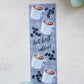 A glossy 2 X 6 bookmark with a pale grey plaid background, depicting cups of coffee and coffee beans with the words “…but first, coffee!” in script lettering across the middle.