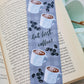 A glossy 2 X 6 bookmark with a pale grey plaid background, depicting cups of coffee and coffee beans with the words “…but first, coffee!” in script lettering across the middle on an open book with leaves in the background.