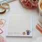 A notepad with a pale yellow grid background with a white border, on the bottom a cup of tea and biscuits are depicted with the words "life begins after chai..." in script lettering. The notepad is on a desk surrounded by pink flowers and stationery supplies as well as a cup of tea.