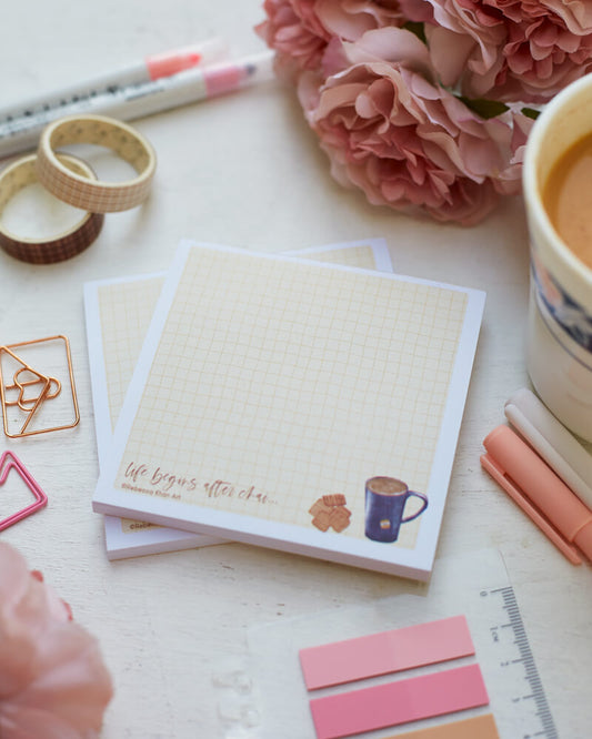Two notepads with a pale yellow grid background with a white border, on the bottom a cup of tea and biscuits are depicted with the words "life begins after chai..." in script lettering.  The notepads are on a desk surrounded by pink flowers and stationery supplies as well as a cup of tea.